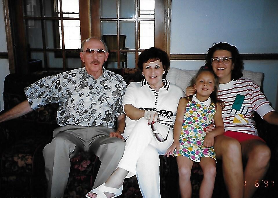 A family of four sits on a dark patterned sofa in a living room, left to right: an older man in a floral shirt, an older woman in white, a young girl in a colorful floral dress, and a woman in a striped shirt with glasses.