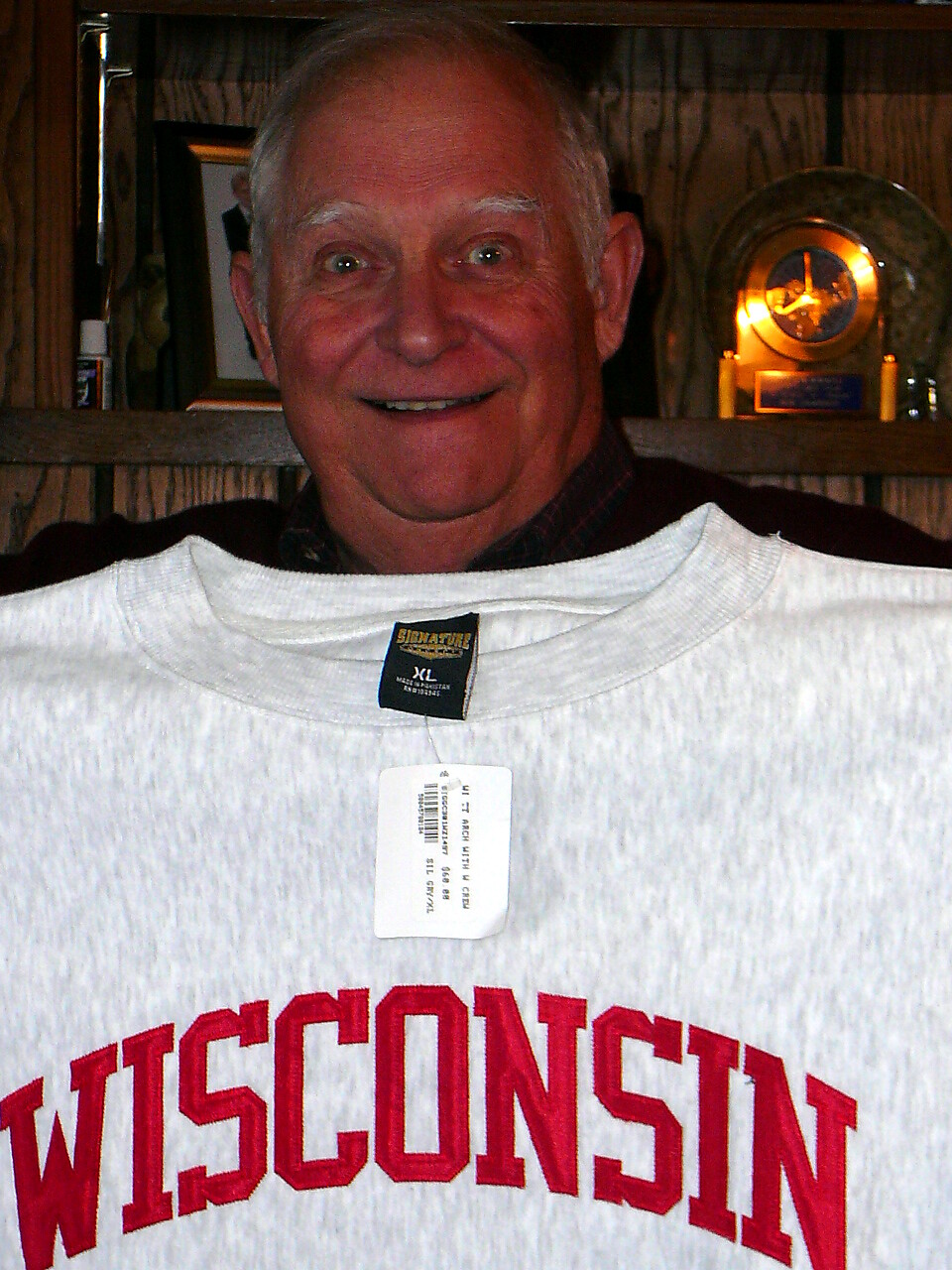 Smiling older man wearing a gray Wisconsin sweatshirt with a price tag hanging from the collar, indoors by wooden shelves and a lamp plaque in the background.
