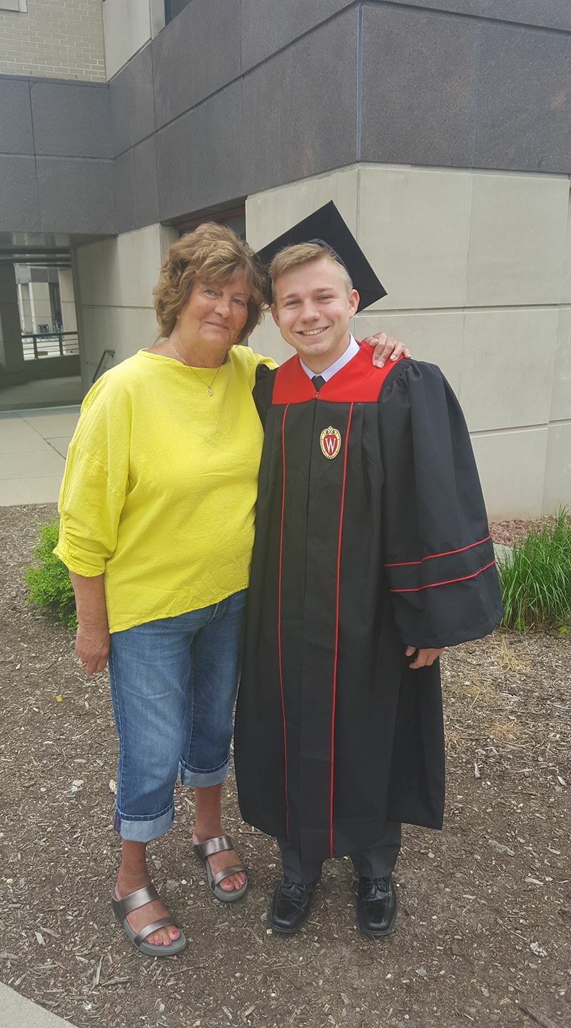 Graduate in black gown with red trim and mortarboard posing with a smiling woman in a yellow top outside a campus-style building. Crest on gown visible.