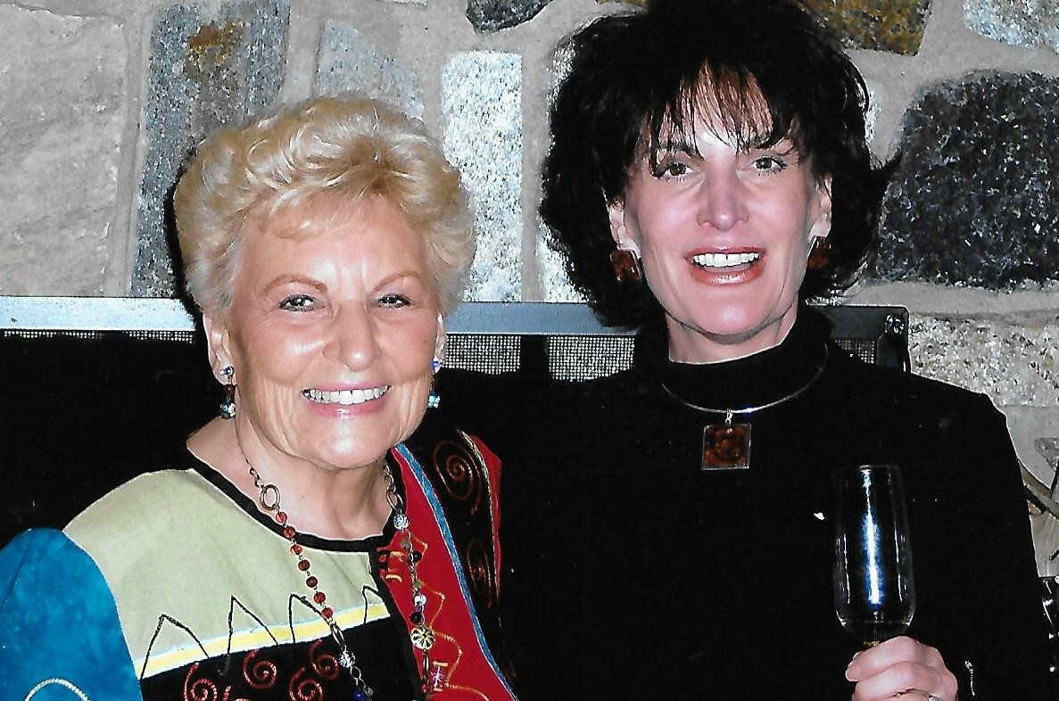 Two smiling women posing together indoors; one wears a colorful patchwork top, the other a black outfit and holds a wine glass.