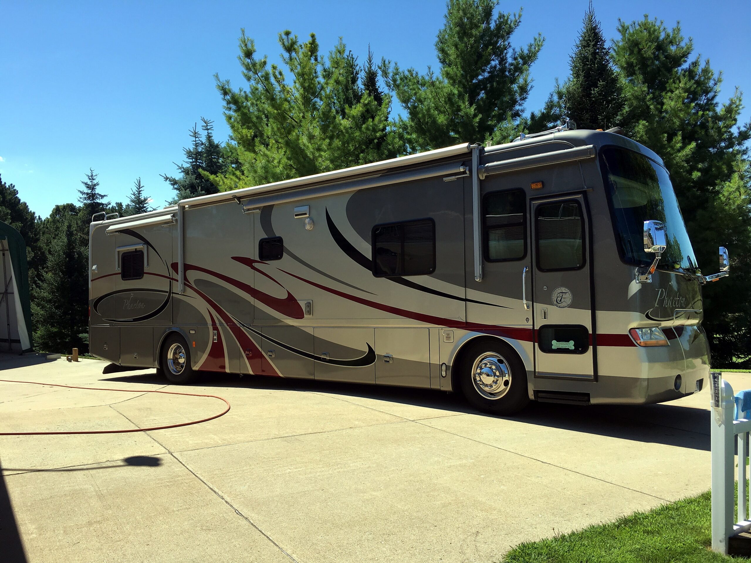 Grey recreational vehicle (RV) parked on a sunny concrete lot with green pine trees and a blue sky in the background.