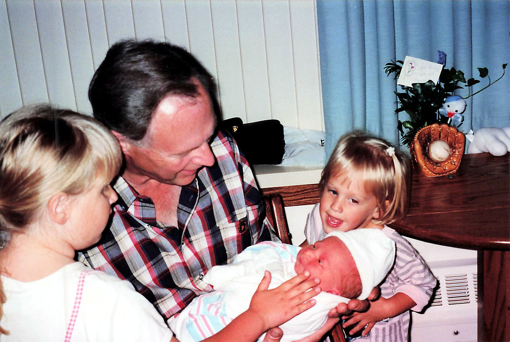 Father in a plaid shirt sits with two young girls, gently cradling a newborn baby wrapped in a blanket in a home setting.