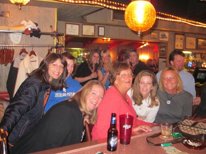 Group of smiling women gathered around a bar counter in a cozy venue, with drinks and a beer bottle on the counter, warm lighting, and framed items on the wall in the background.