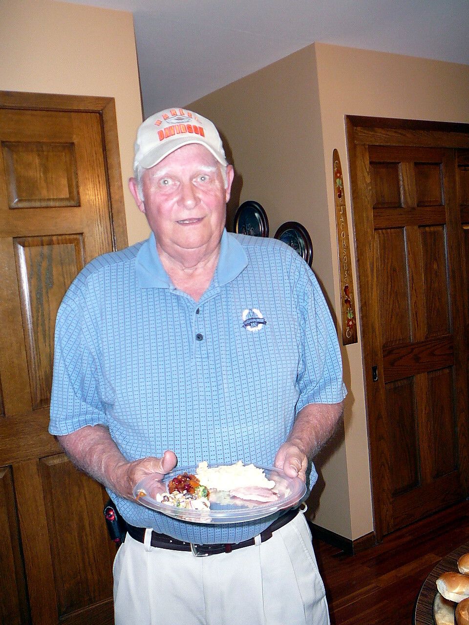 Older man in a light blue polo and white Harley-Davidson cap, holding a plate of food at a home gathering.