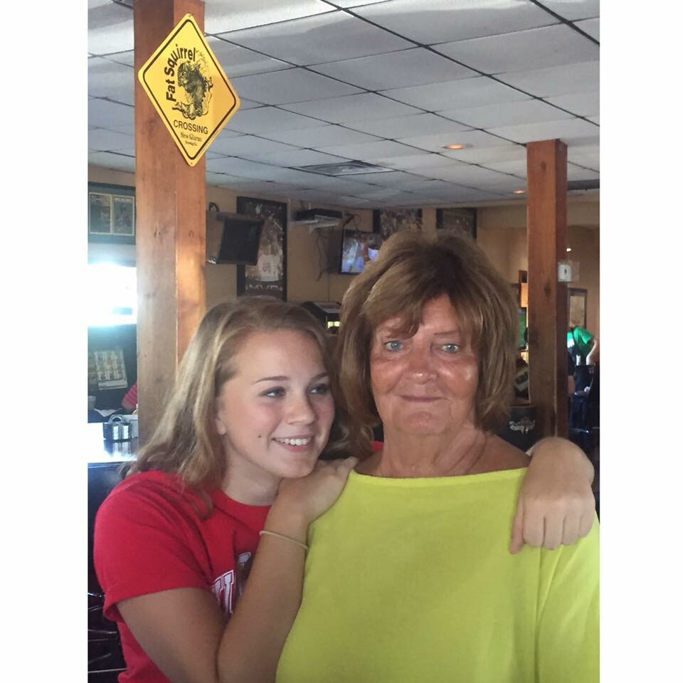 Two women, a younger girl in red and an older woman in a bright yellow top, smile as they hug in a casual restaurant; a yellow 'Fat Squirrel Crossing' sign is on a wooden pillar behind them.