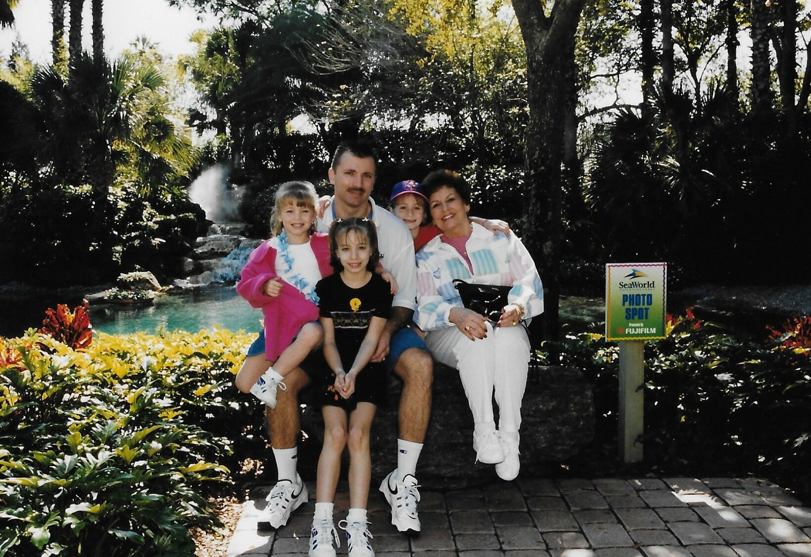 Family of five posing on a stone bench at SeaWorld Photo Spot, with a water feature and tropical plants behind them.