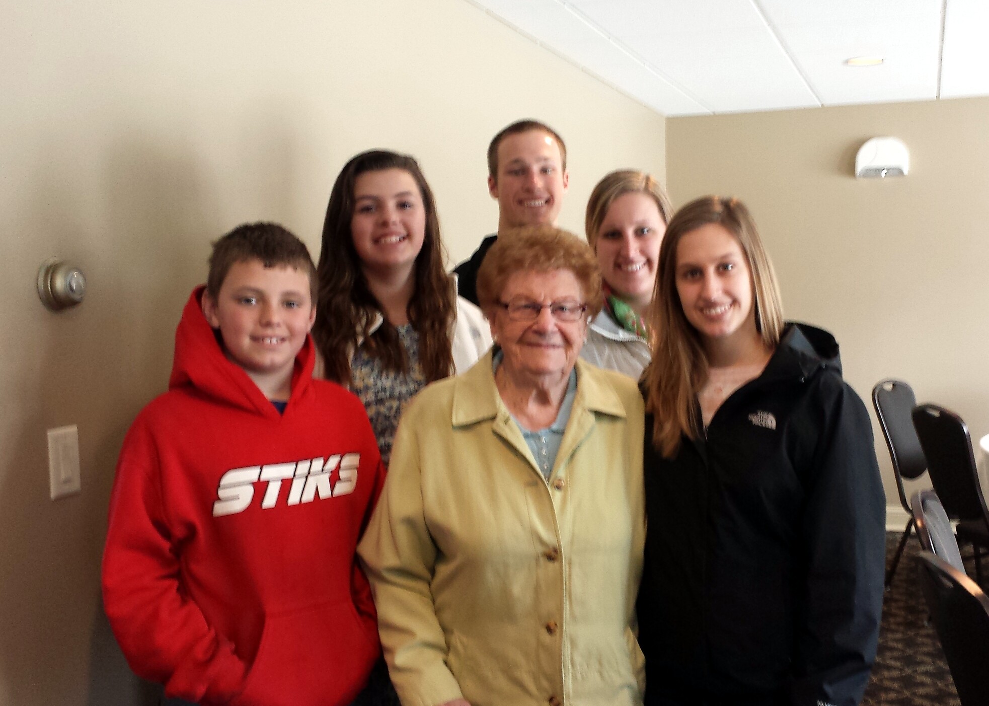 Group of five people smiling for a photo indoors, with an elderly woman in a beige coat at the center.