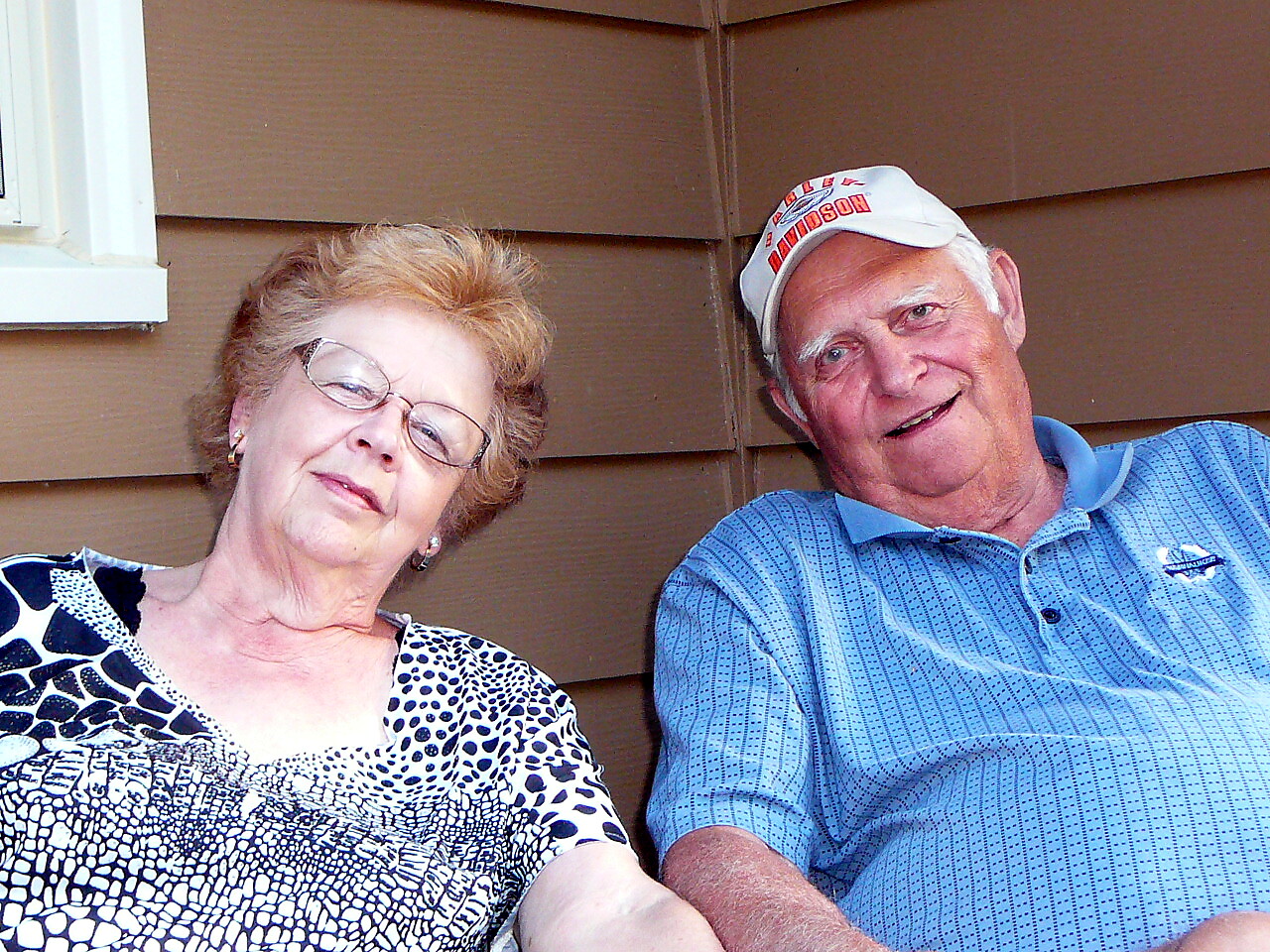 Senior couple sitting side by side outside, smiling at the camera; woman in patterned blouse with glasses, man in blue polo and white cap.
