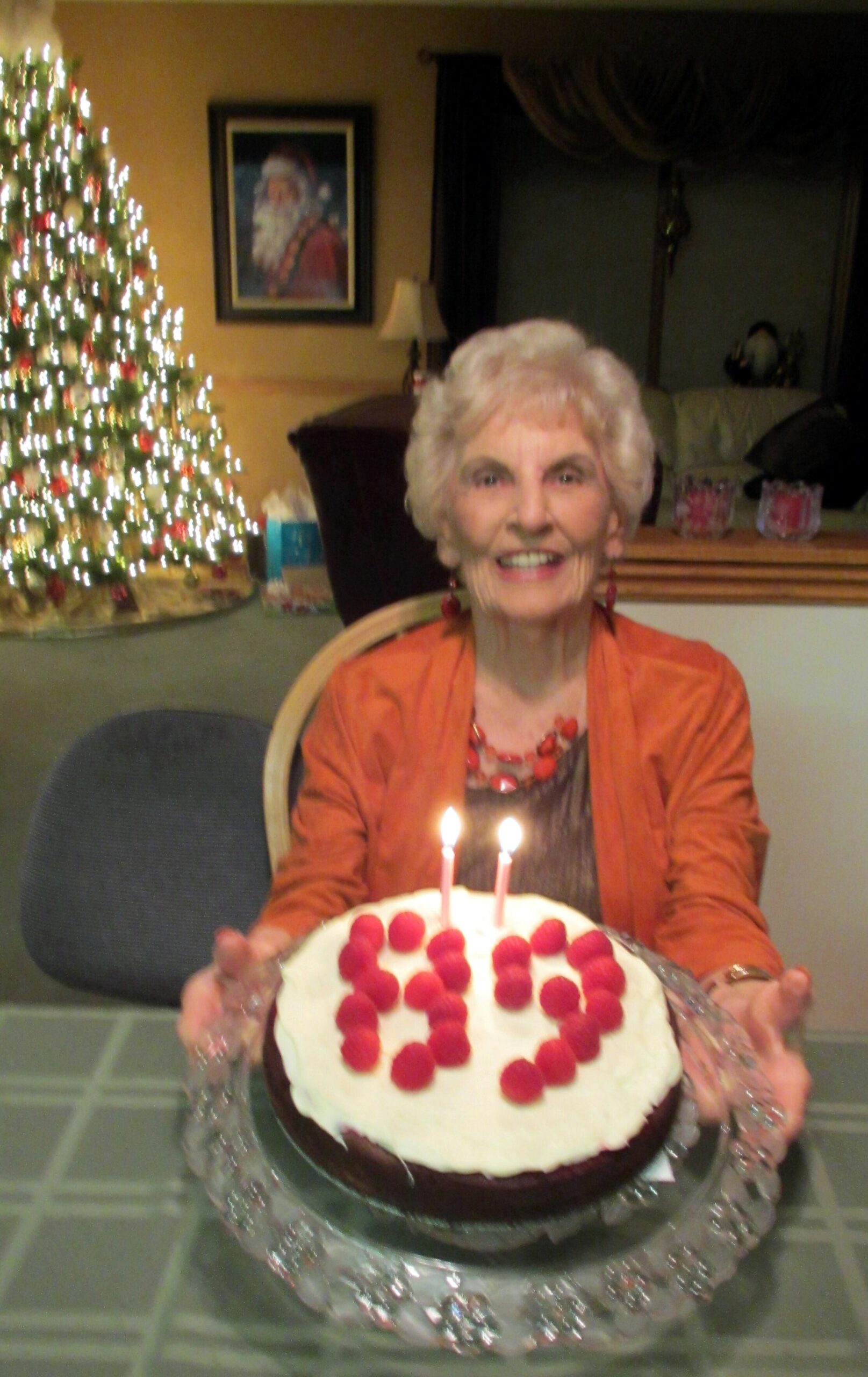 Smiling elderly woman in orange jacket presents a white-frosted cake with red raspberries and two candles, in a decorated room with a Christmas tree in the background.