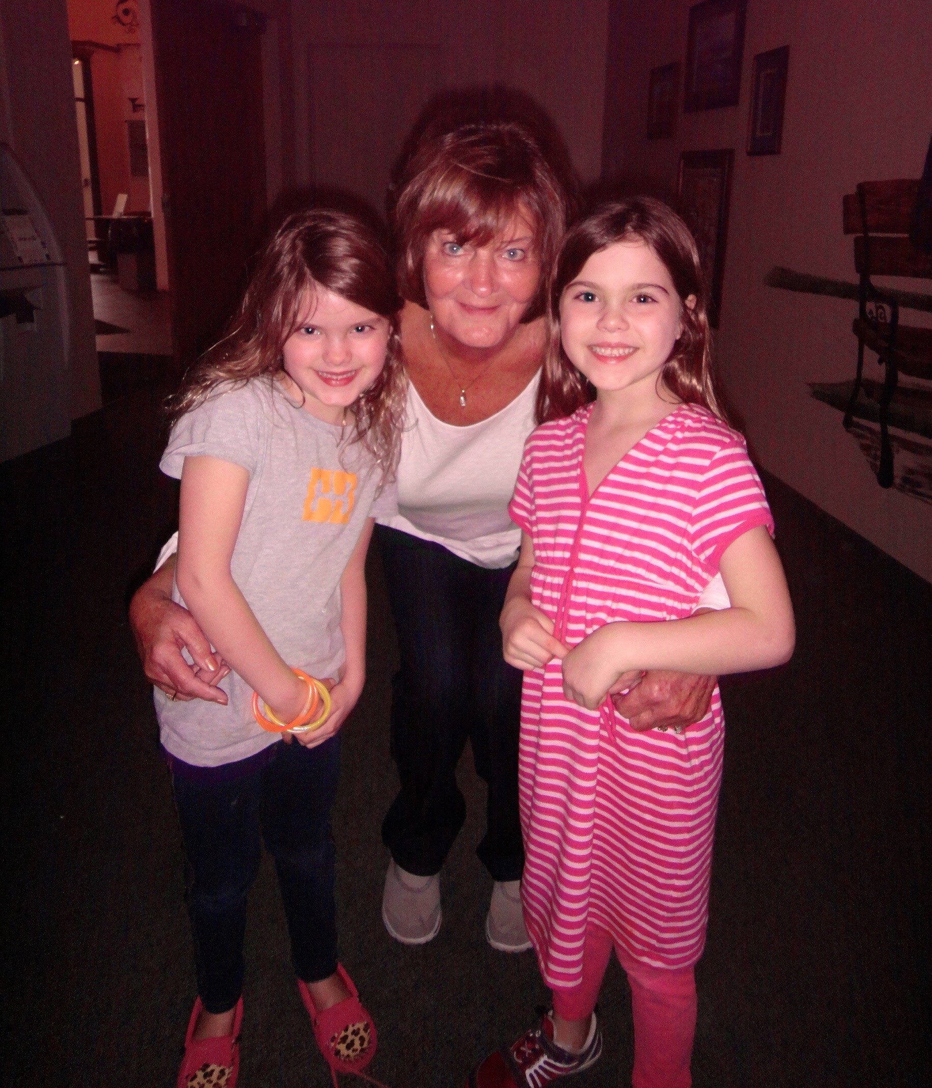 Three generations posing together: an elderly woman flanked by two smiling young girls indoors. The girl on the left wears a gray t-shirt and bright bracelets; the girl on the right wears a pink striped dress and pink leggings.