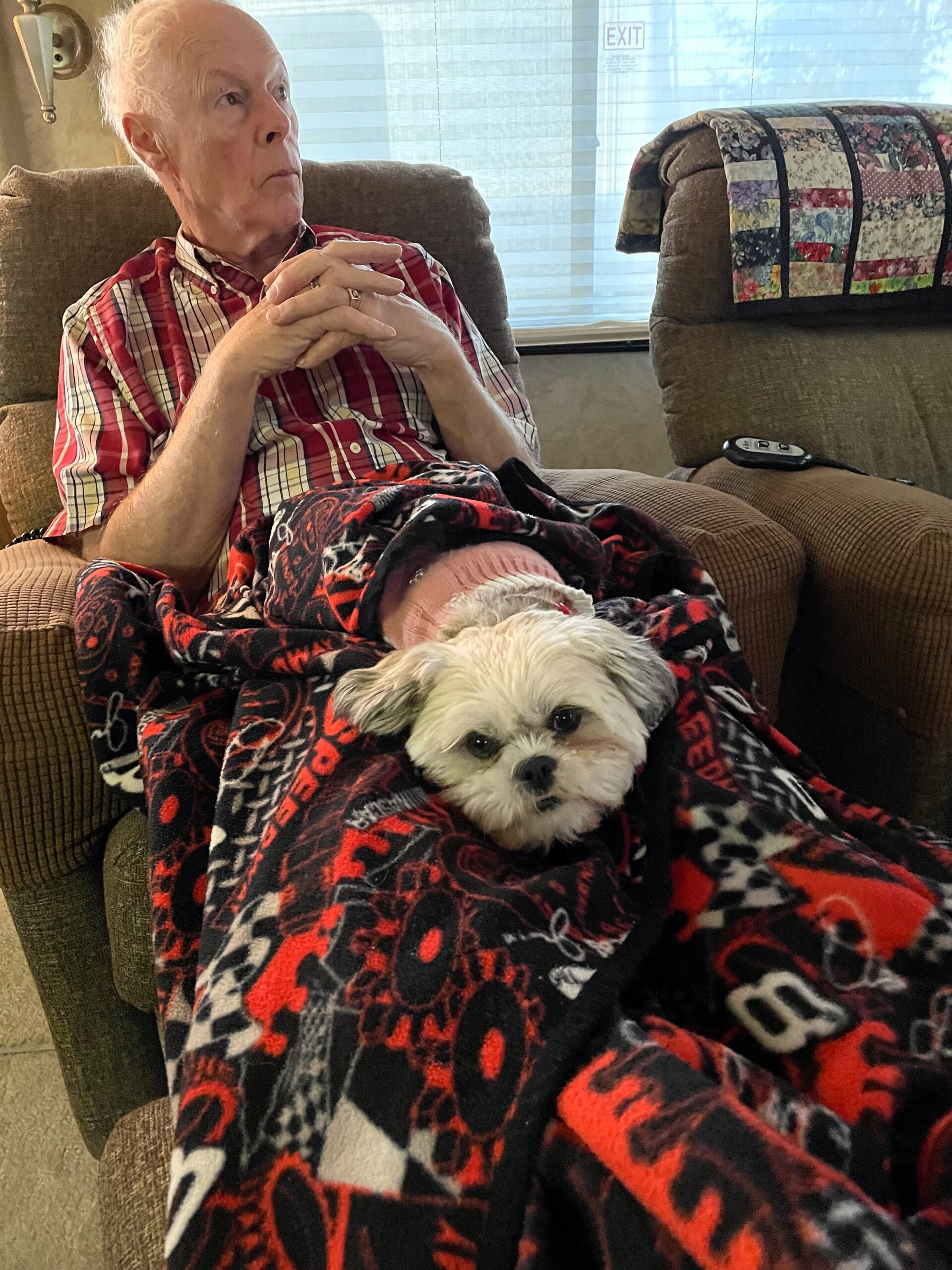 An elderly man in a red plaid shirt sits in a brown recliner, hands clasped, with a small white dog on his lap under a red and black blanket.