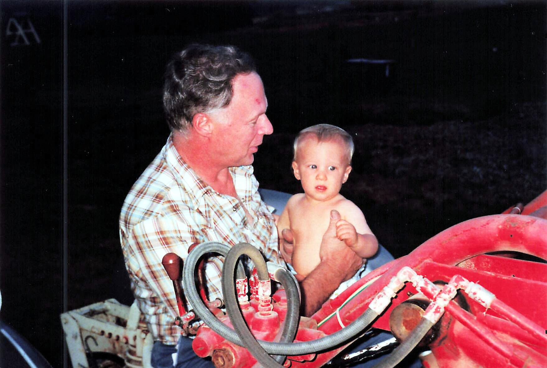 Older man in a plaid shirt holds a baby on a red farm machine at night.