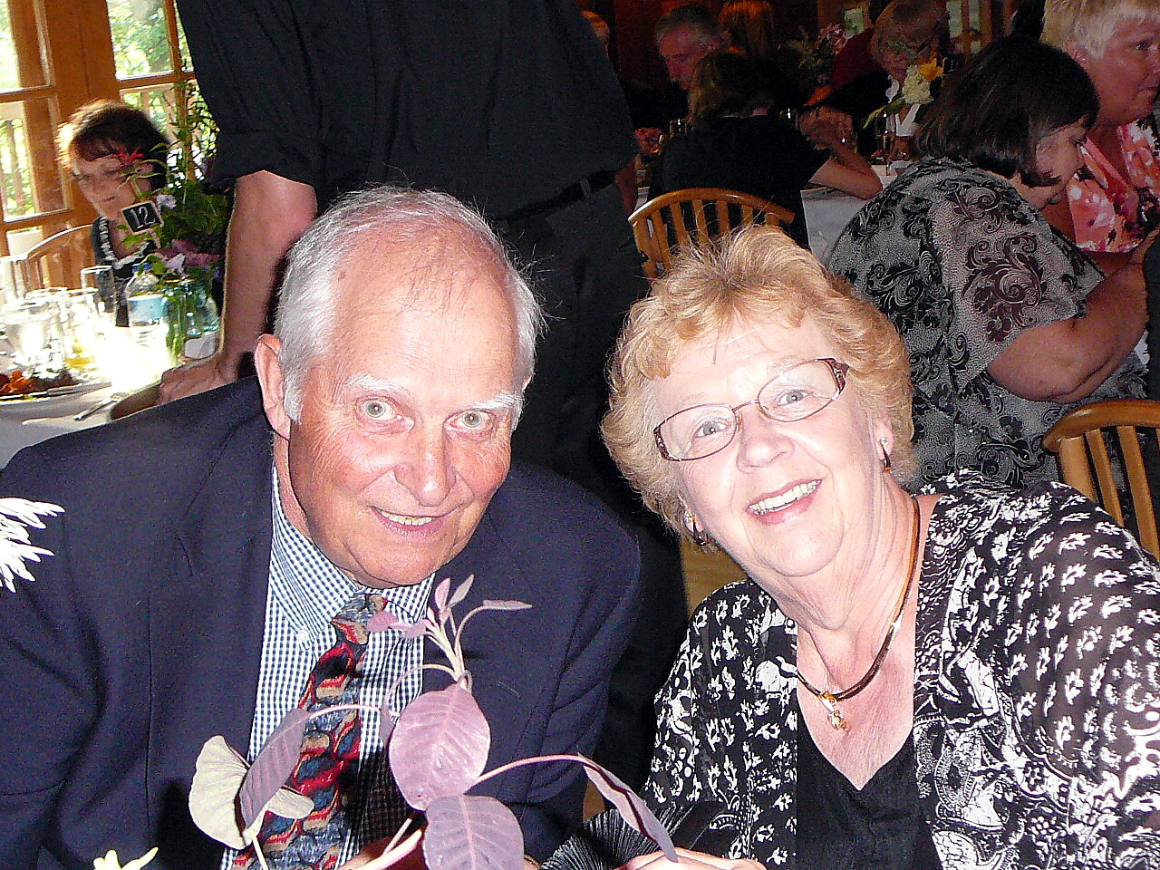 Smiling older couple at a table during a formal event or wedding reception, with flowers and guests in the background.