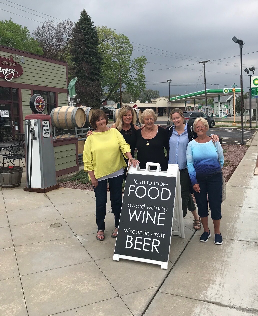 Five women pose beside a chalkboard sign advertising farm-to-table food, wine, and Wisconsin craft beer outside a winery.