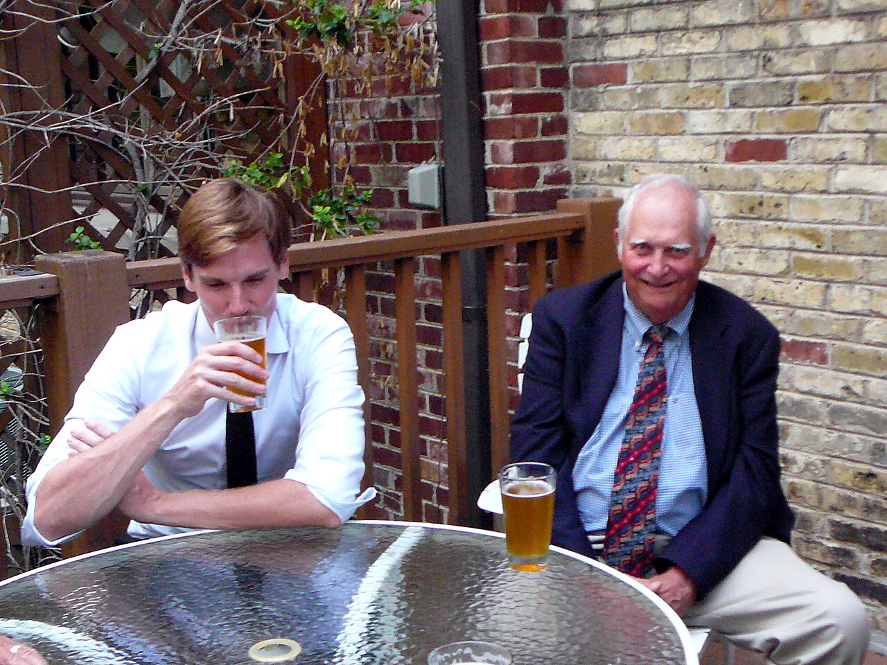 Two men sit at a glass outdoor table with beers; the younger man in a white shirt and black tie sips his drink, while the older man in a blazer smiles nearby.