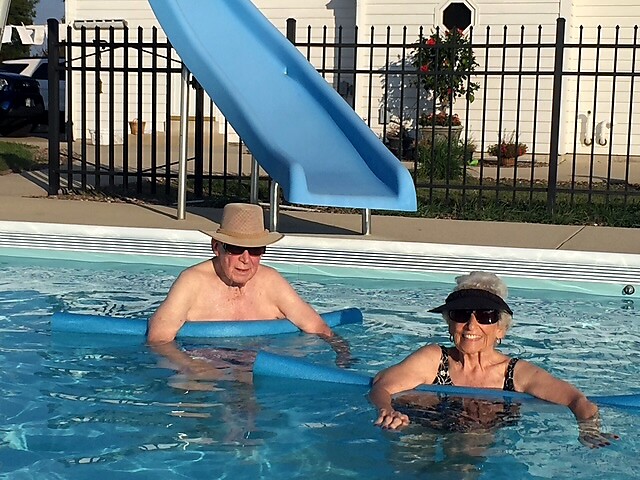 Two seniors enjoy a sunny pool day, each holding a blue pool noodle while leaning on the water’s surface with a safety fence and a blue slide in the background.