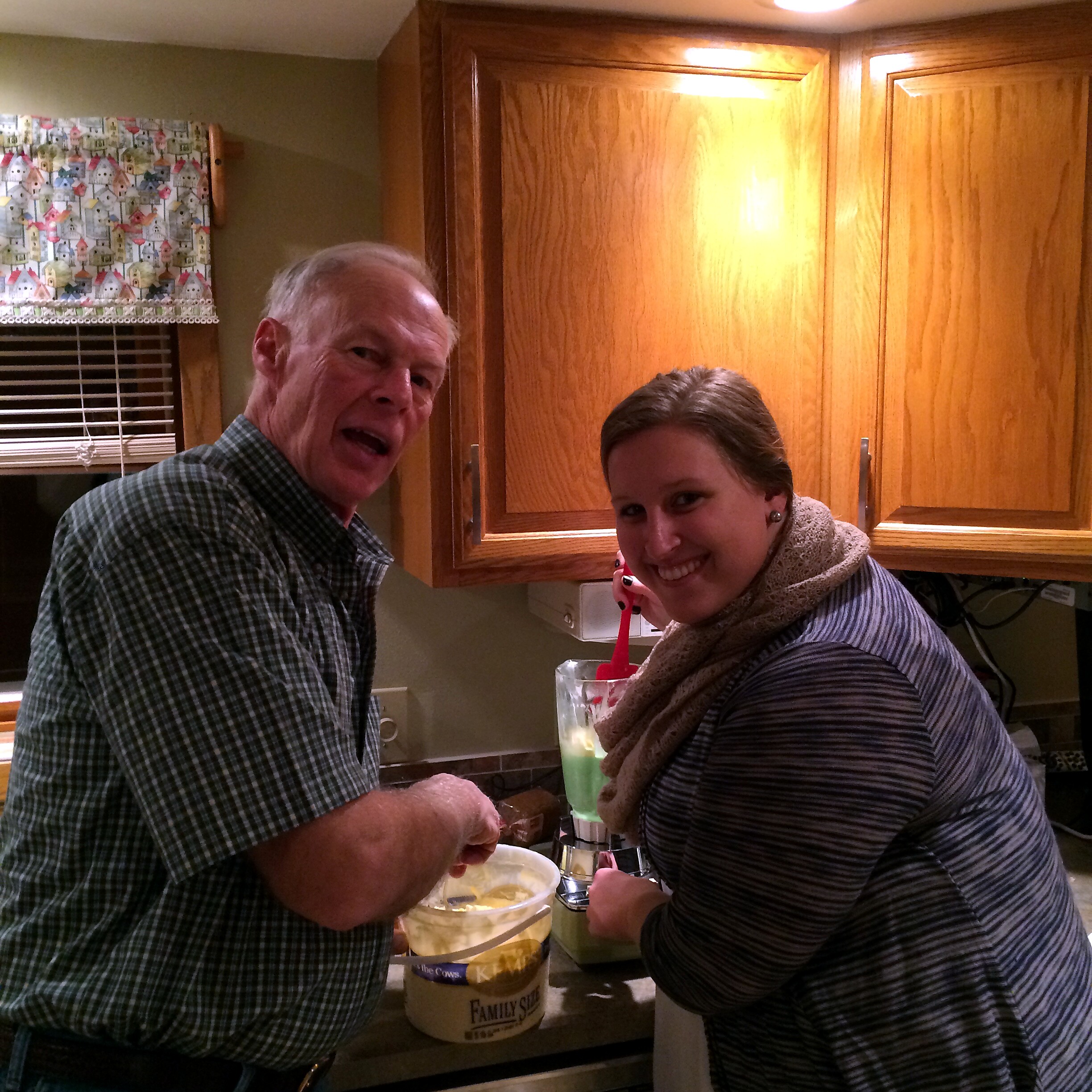Two people in a kitchen scoop ice cream from a large tub labeled Family Size, with wooden cabinets in the background.