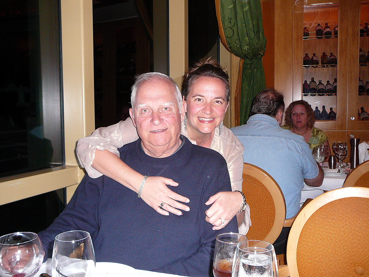 A smiling couple sits at a restaurant table; the woman hugs the man from behind, both looking at the camera.