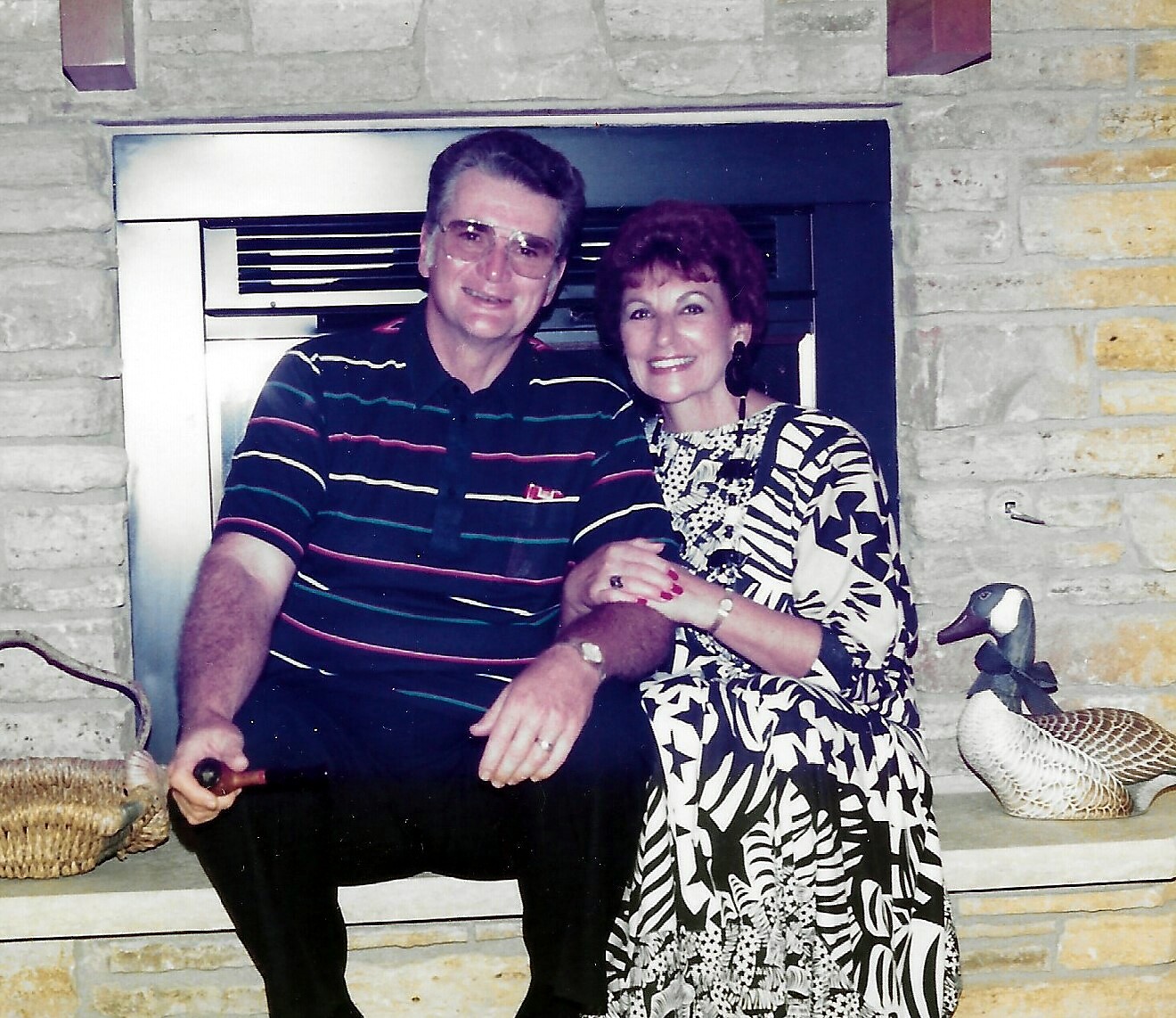 Smiling older couple seated on a stone fireplace hearth, man in striped polo and glasses, woman in a black-and-white patterned dress; decorative ducks nearby.