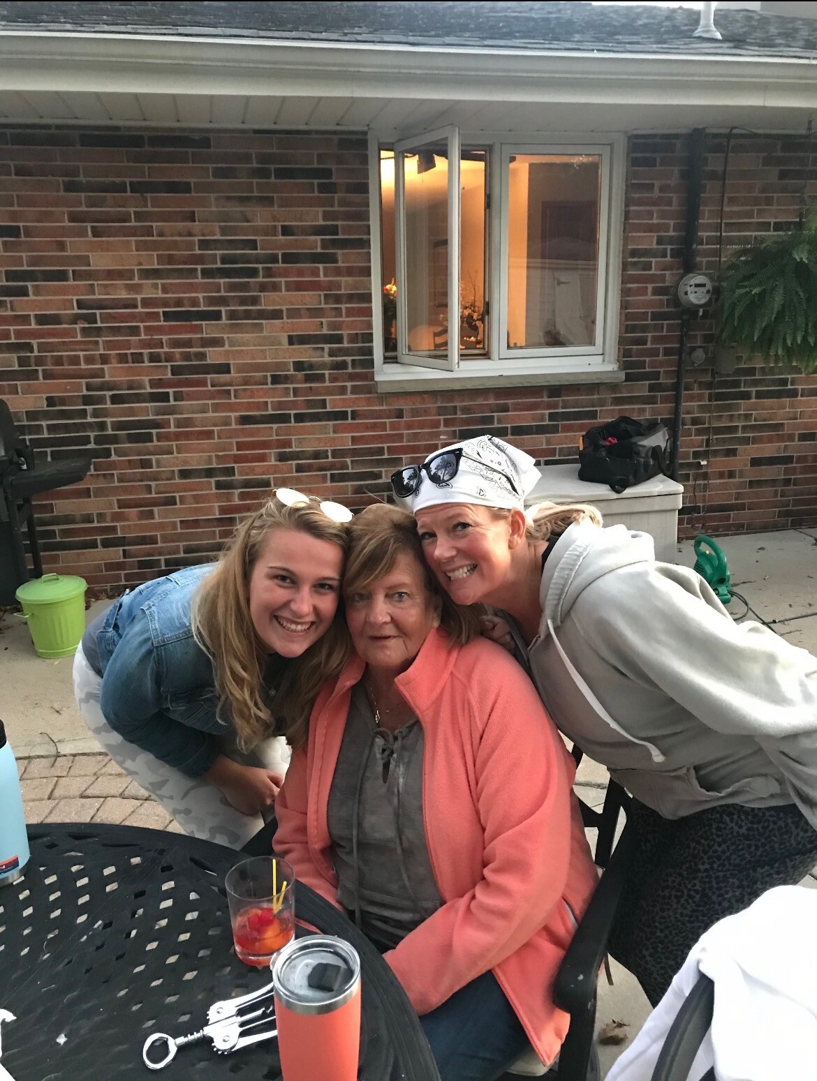 Three generations posing together at an outdoor patio, smiling at the camera with a brick house in the background.