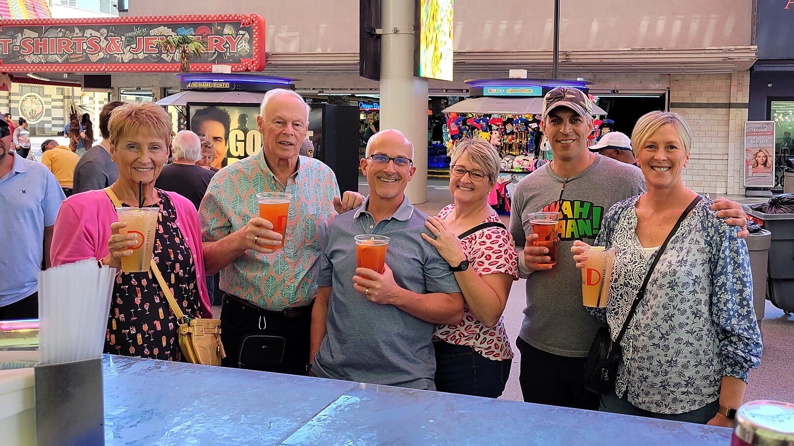 Six adults pose with drinks at an outdoor street fair, smiling for the camera.