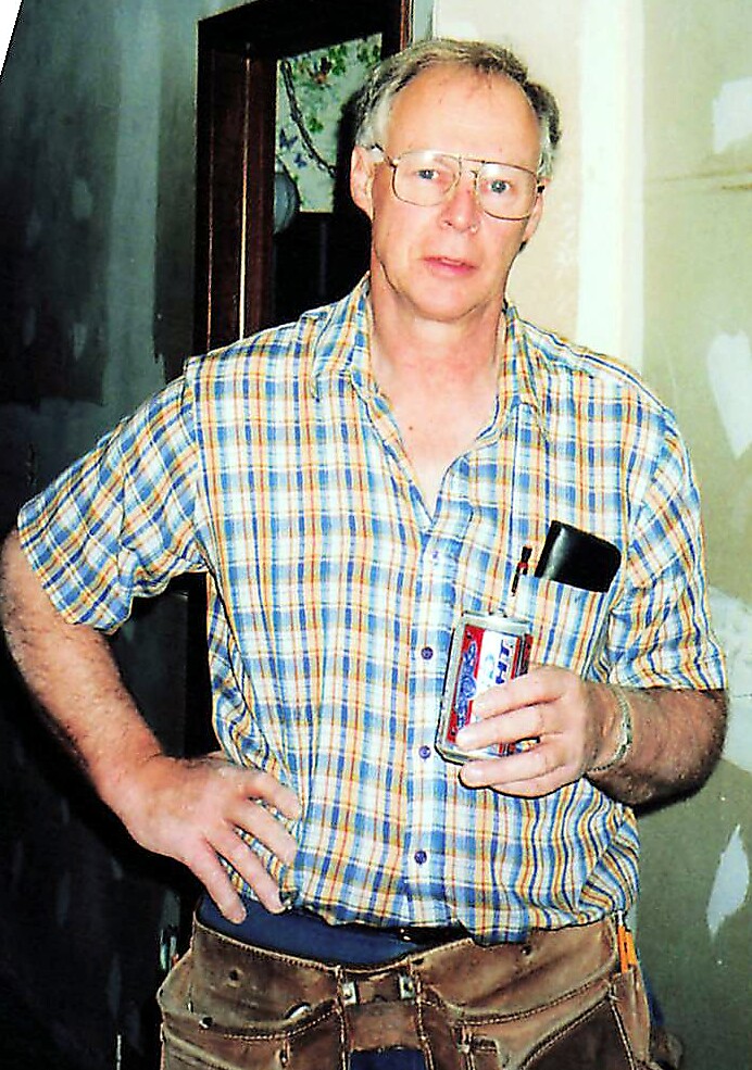 Middle-aged man in a plaid shirt and tool belt stands indoors with one hand on his hip, holding a beverage can and a phone in his pocket.