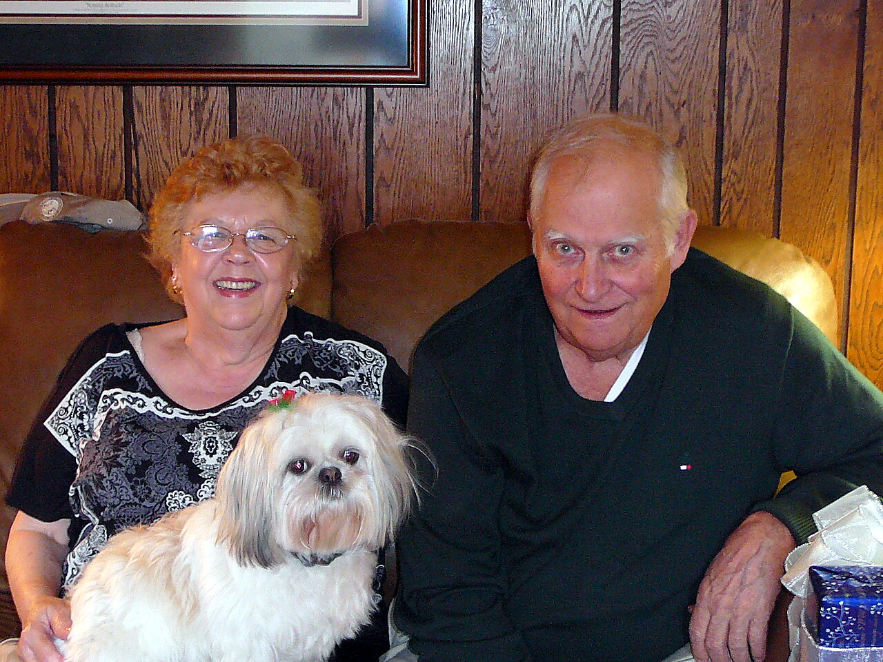 Smiling elderly woman and man sitting on a brown leather couch with a fluffy white dog in front of them.