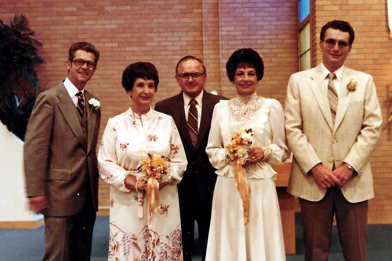 Five people pose indoors at a wedding: two brides in white gowns with bouquets, flanked by two men in beige suits and a man in a brown suit.