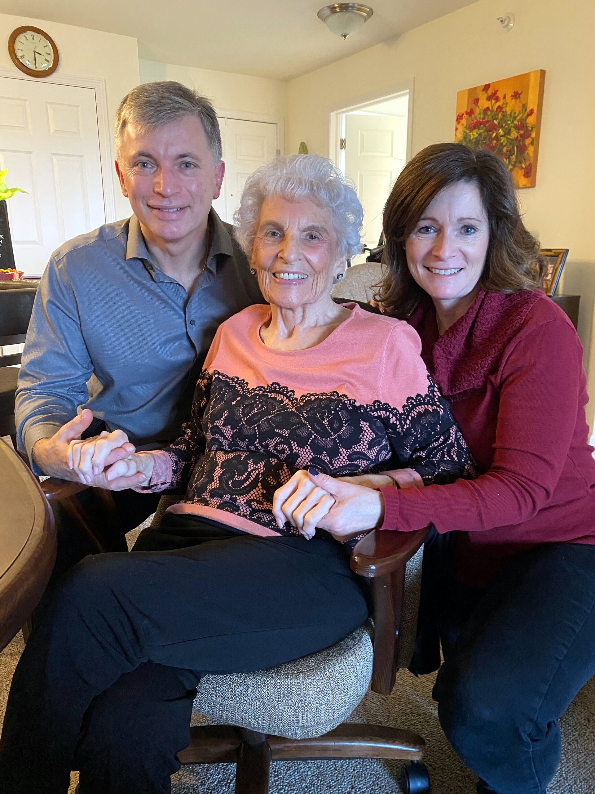 Three people sit close together indoors, smiling at the camera; an elderly woman is flanked by a man and a younger woman, hands joined in front.
