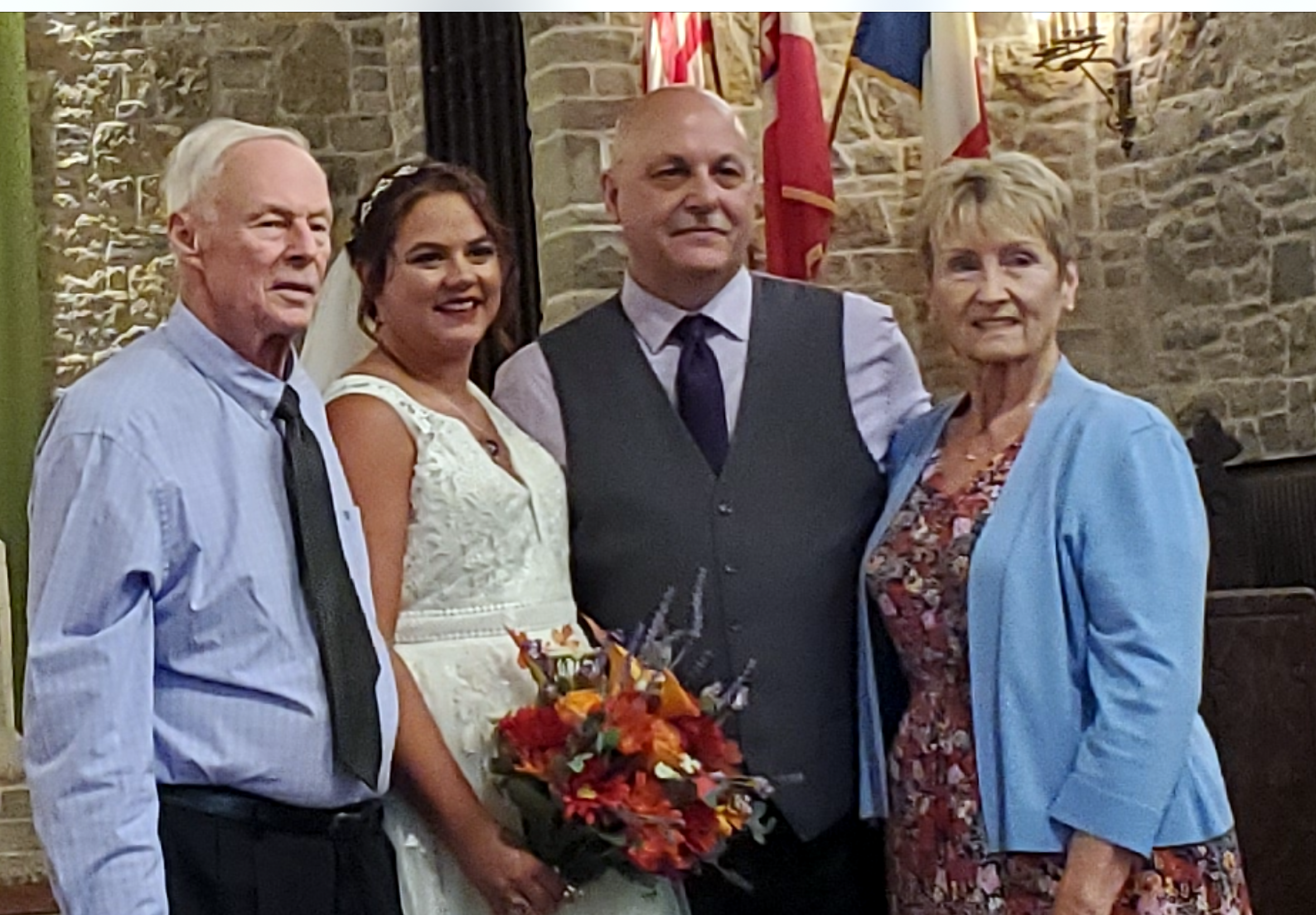 Group of four adults posing for a wedding photo; bride in white dress holding a bouquet of orange flowers, flanked by two men in vests and a woman in a blue cardigan.