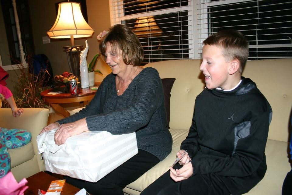 Woman and a boy on a couch opening a wrapped gift together during a casual gathering, warm lamp light in the background.