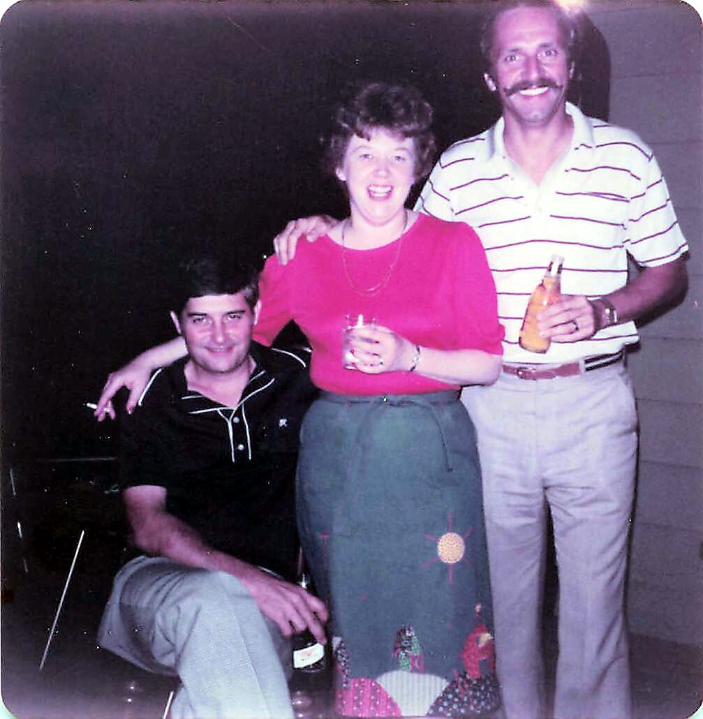 Three adults posing for a casual group photo: a woman in a pink top and patterned skirt between two men, each holding a drink, smiling at the camera.