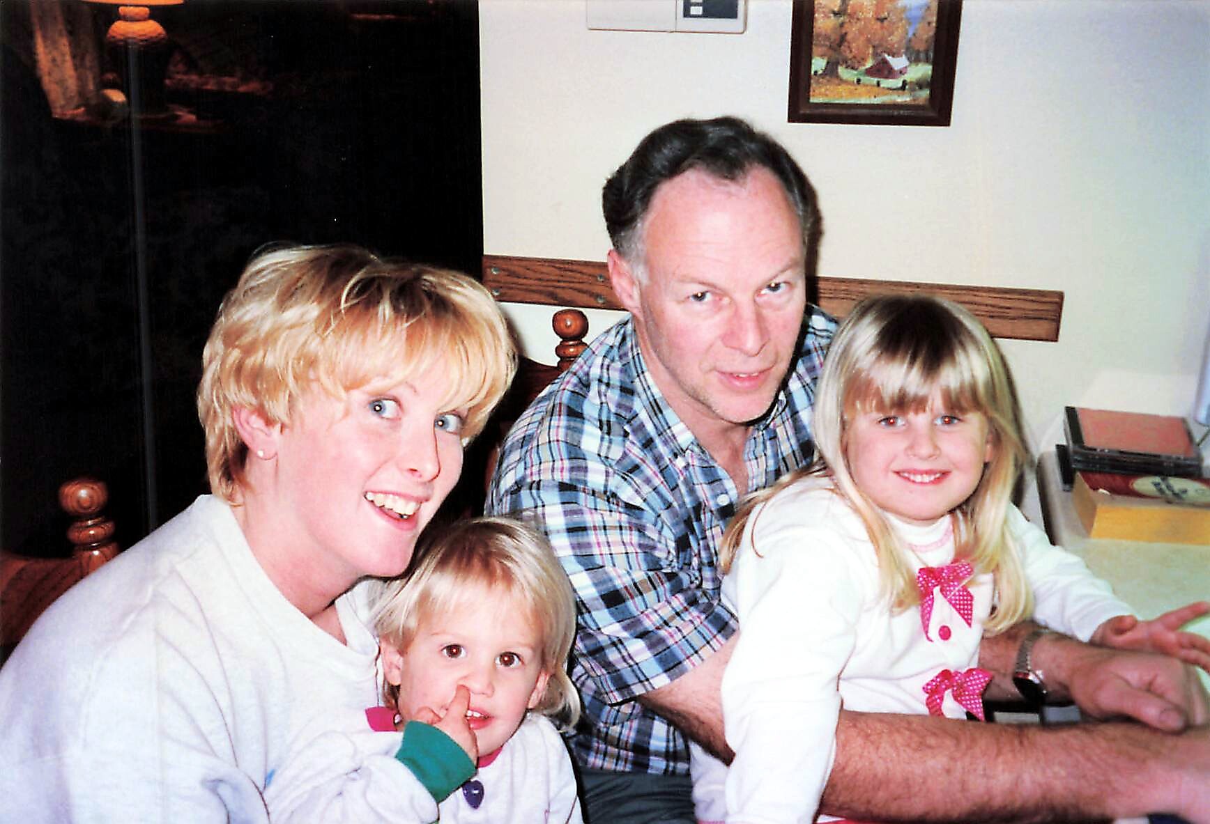 A smiling family at a table: woman on left, man in plaid shirt, and two young girls closer to the camera indoors.