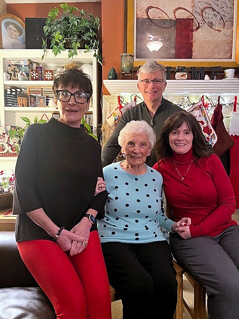 A group of three generations posing together in a cozy festive living room, smiling at the camera, with holiday stockings in the background.