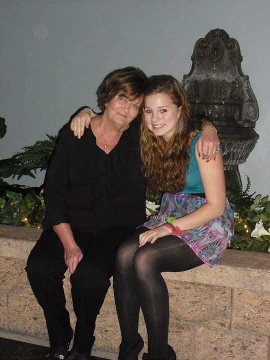 Two women sit close together on a stone ledge, smiling; older woman in black and younger woman in teal top with a patterned skirt, with a fountain in the background.