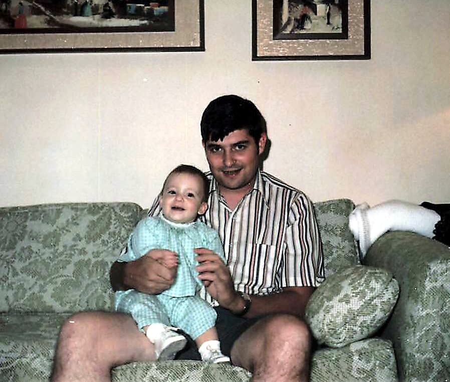 Man in a striped shirt sits on a green patterned couch holding a smiling baby in a light blue outfit.