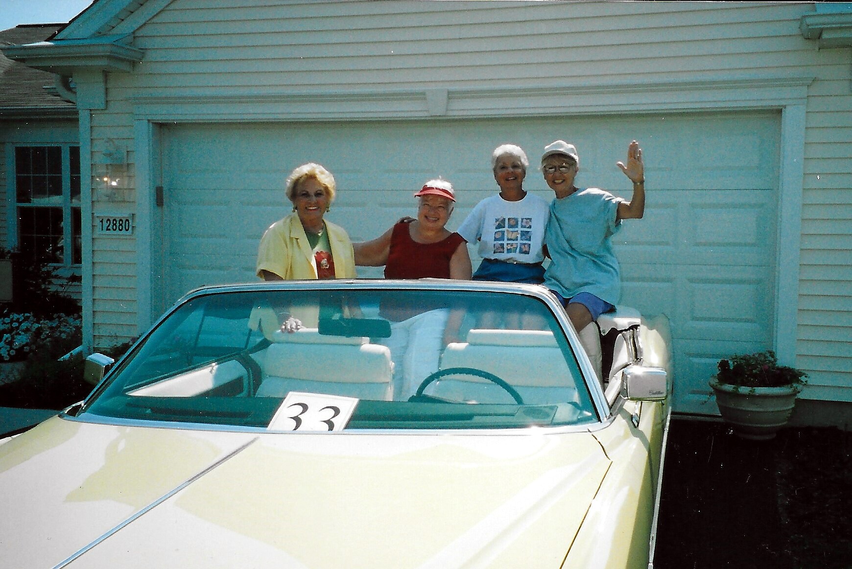 Four smiling adults sit on a light-yellow convertible in a driveway, posing for the photo and waving.