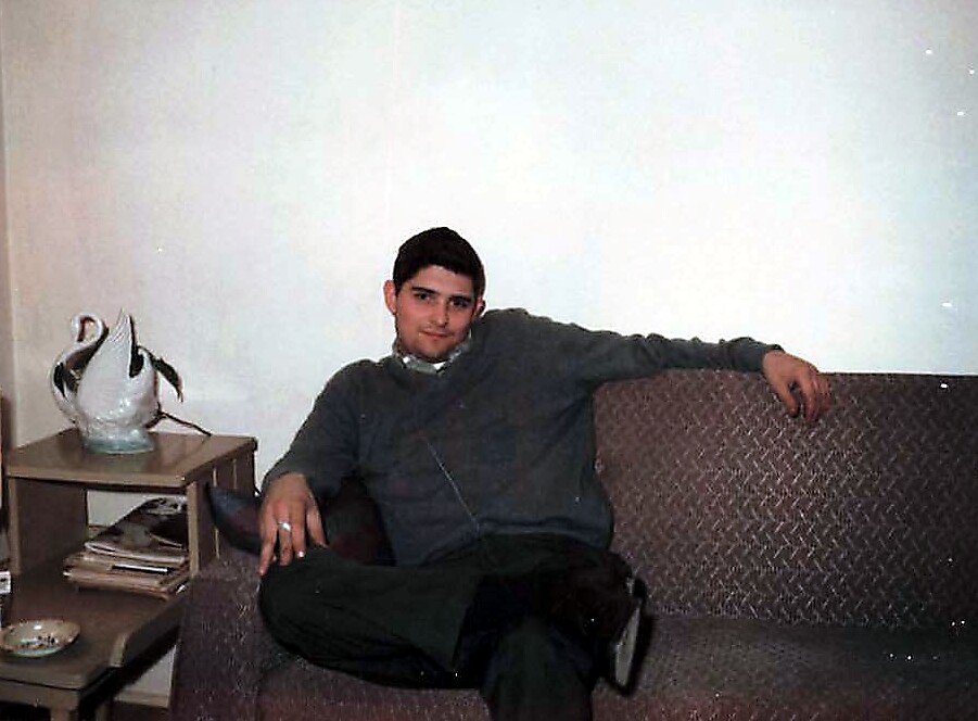 Man seated on a patterned couch with legs crossed, wearing a gray sweater and headphones, a swan figurine on a side table nearby and magazines underneath it