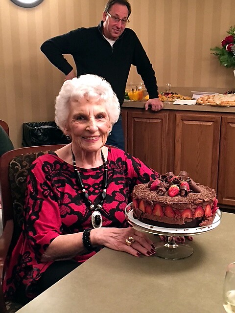 Smiling elderly woman in a pink and black top sits at a table, with a chocolate-strawberry cake on a stand in front; a man stands behind near a buffet table.