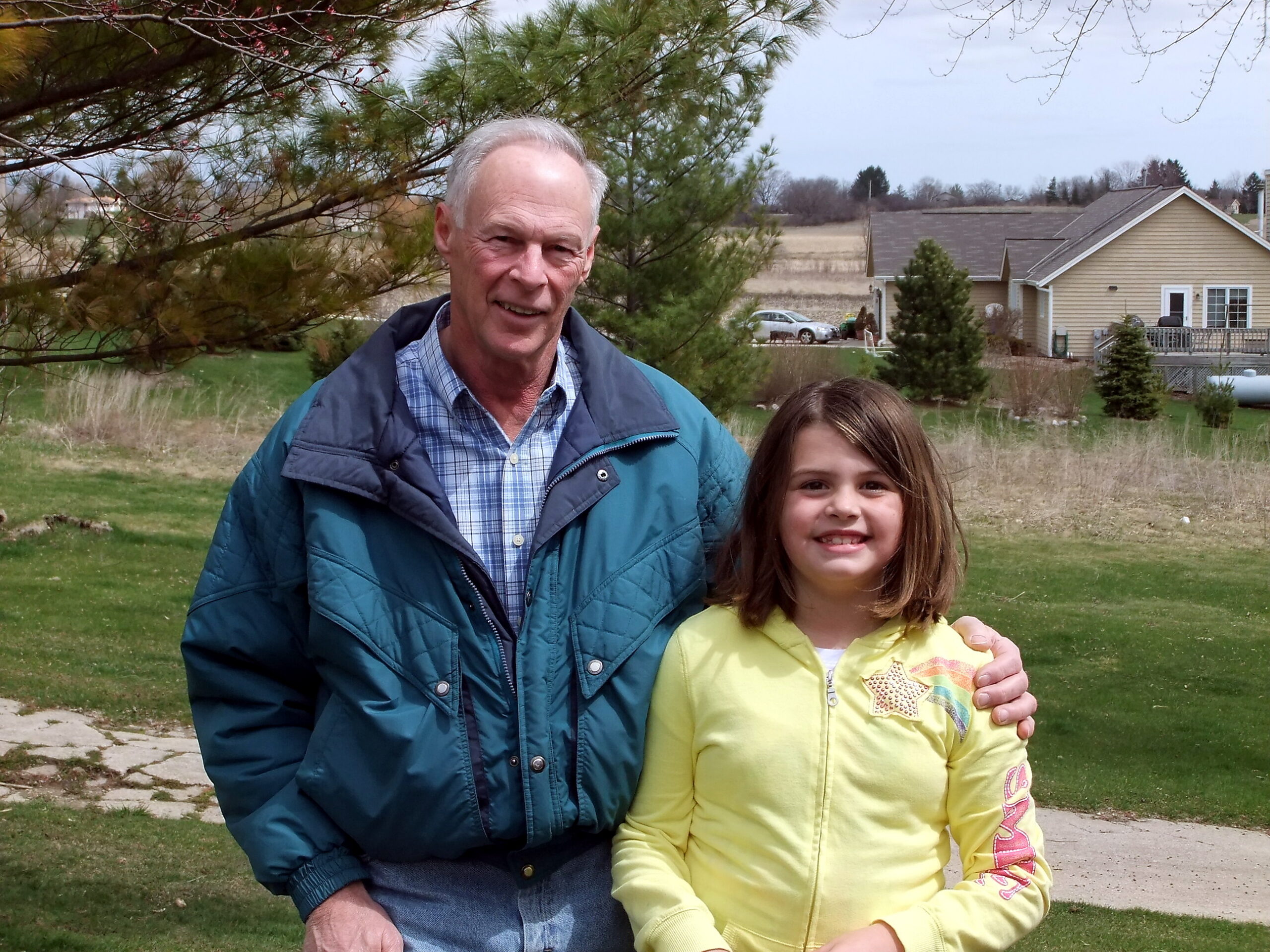 Two people, an older man and a young girl, posing together outdoors with trees and a house in the background, smiling at the camera.