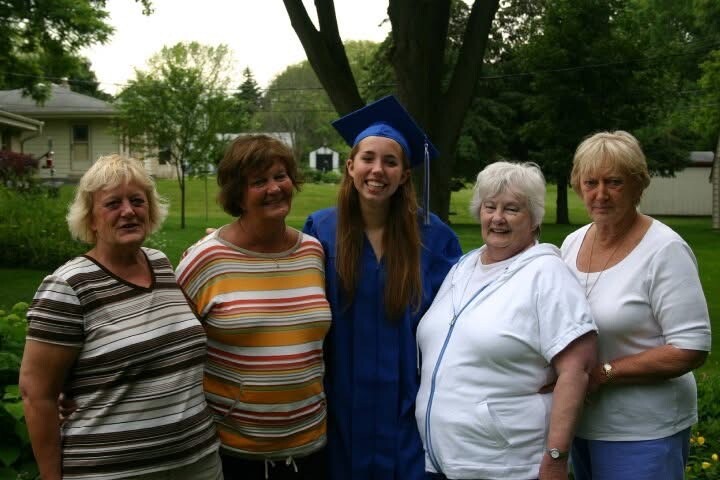 Graduate in blue cap and gown posing with four women outdoors in a park-like yard, celebrating on graduation day.