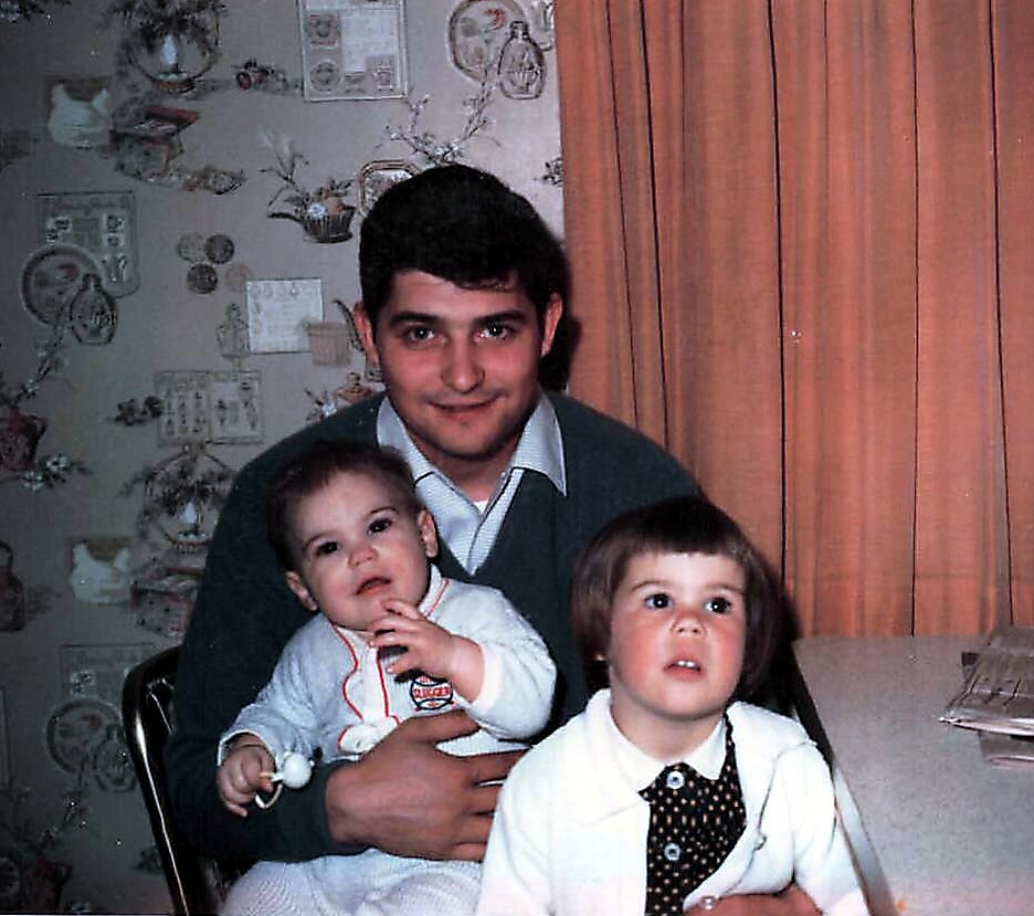 Man with dark hair holding a baby and sitting with a young girl indoors, all looking toward the camera.