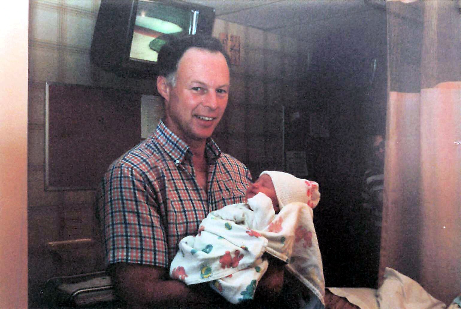 Smiling man in a plaid shirt holding a newborn baby wrapped in blankets in a hospital room.