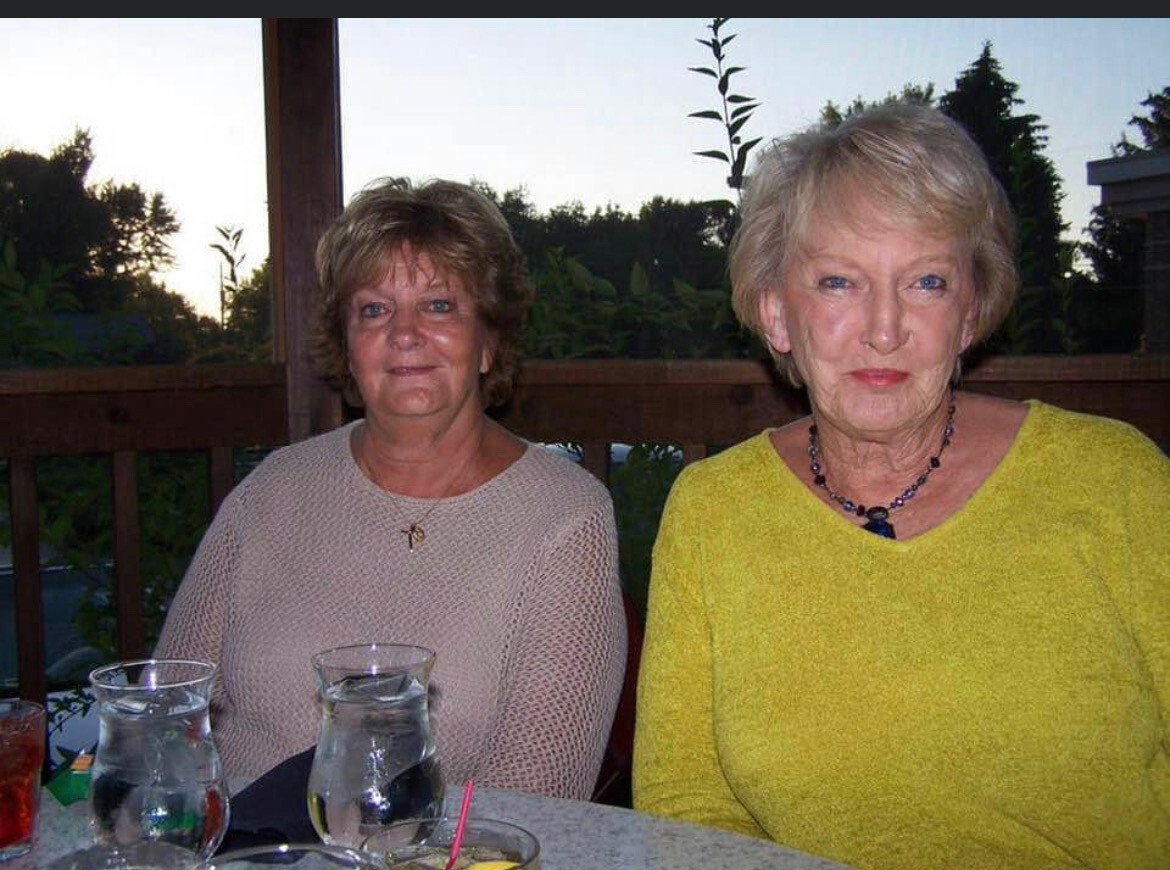 Two older women sit at an outdoor deck table with drinks, smiling at the camera as evening light fades behind them.