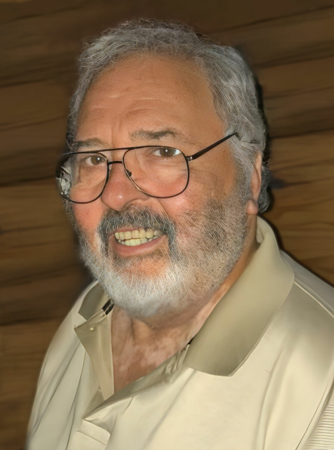Smiling older man with gray hair and beard wearing glasses and a light beige shirt, looking at the camera, indoors.