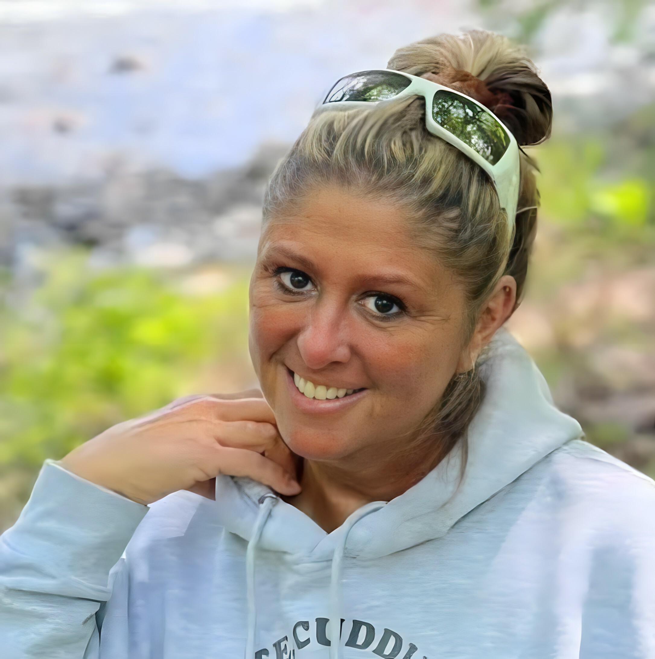 Woman smiling at the camera with white sunglasses perched on her head outdoors.