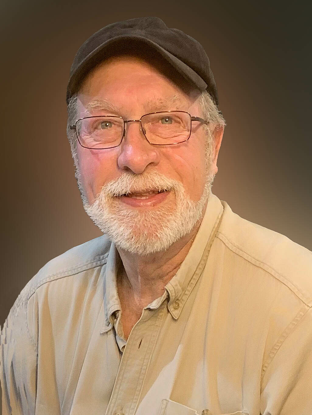 Portrait of a smiling older man with a white beard, glasses and a black cap against a neutral background.