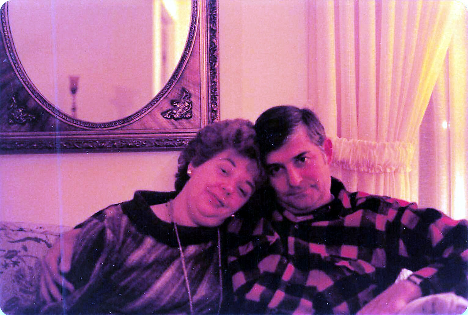 Smiling couple sitting close together on a sofa, heads touching, in a living room with a decorative mirror behind them.