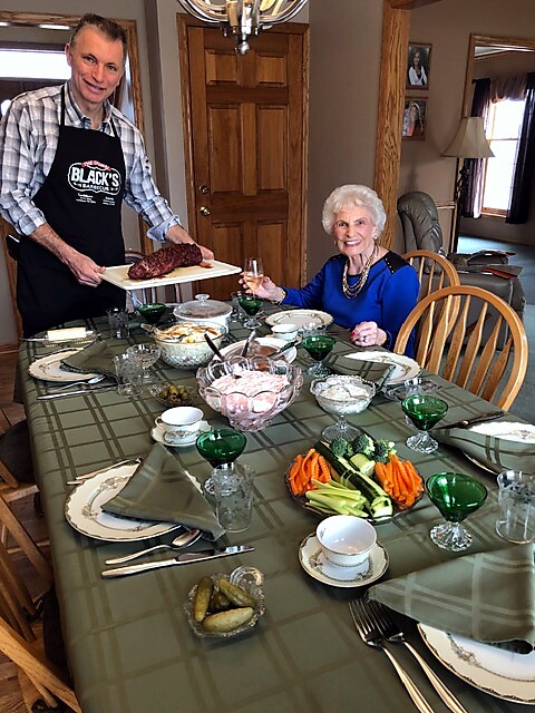 Man in a plaid shirt and apron serves roast meat to an elderly woman at a formal home dining table; she toasts with a glass.
