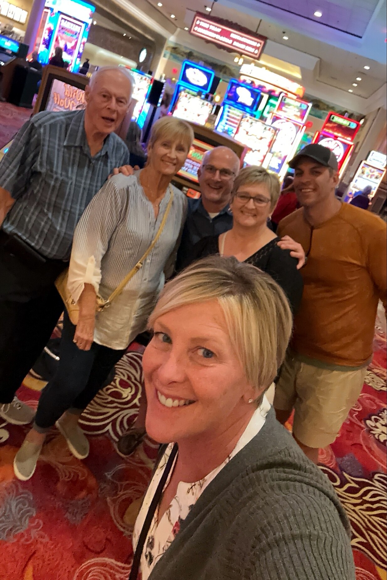 Smiling woman in the foreground taking a selfie with friends on a casino floor filled with colorful slot machines behind them.